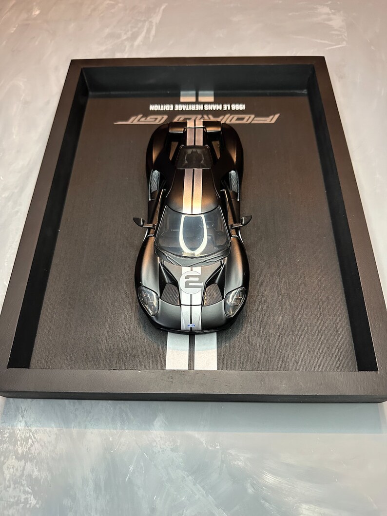 May include: A black and silver Ford GT model car, displayed in a black tray. The car has white racing stripes and the number 2 on the hood. The tray has the text "1966 Le Mans Heritage Edition" and "Ford GT".