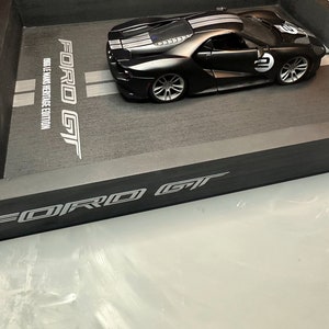 May include: A black Ford GT model car with white racing stripes and the number 2 on the side, displayed in a black tray. The tray has the words "FORD GT" and "1966 LE MANS HERITAGE EDITION" printed on it.