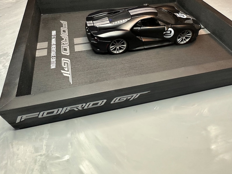 May include: A black Ford GT model car with white racing stripes and the number 2 on the side, displayed in a black tray. The tray has the words "FORD GT" and "1966 LE MANS HERITAGE EDITION" printed on it.
