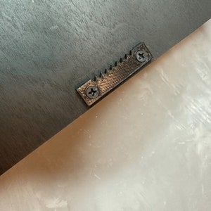 May include: Close-up of the back of a black wooden frame. A metal sawtooth hanger is attached with two screws. The hanger is centered on the frame and is designed for hanging the frame on a wall.