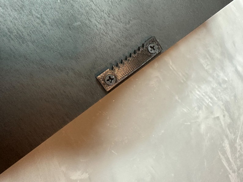 May include: Close-up of the back of a black wooden frame. A metal sawtooth hanger is attached with two screws. The hanger is centered on the frame and is designed for hanging the frame on a wall.