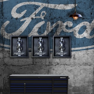 May include: Three framed Ford GT model car displays hang on a textured wall with a large Ford logo. The displays are above a black and blue tool chest. A vintage-style light fixture hangs from the ceiling.
