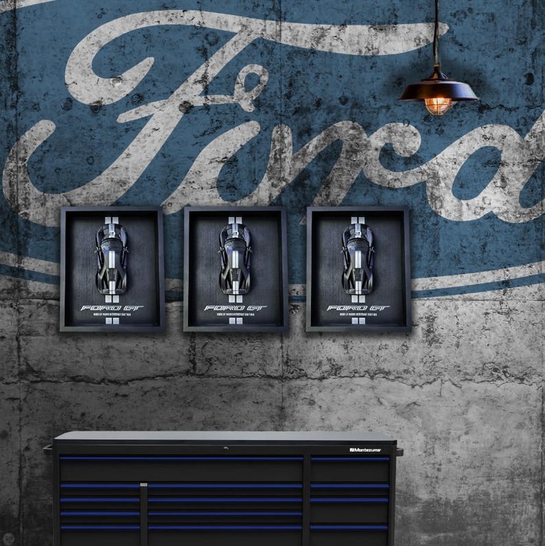 May include: Three framed Ford GT model car displays hang on a textured wall with a large Ford logo. The displays are above a black and blue tool chest. A vintage-style light fixture hangs from the ceiling.
