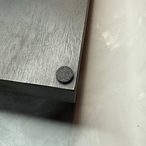 May include: A corner view of a gray wooden board with a dark gray felt pad attached to the bottom corner. The wood grain is visible on the surface of the board. The background is a light, textured surface.