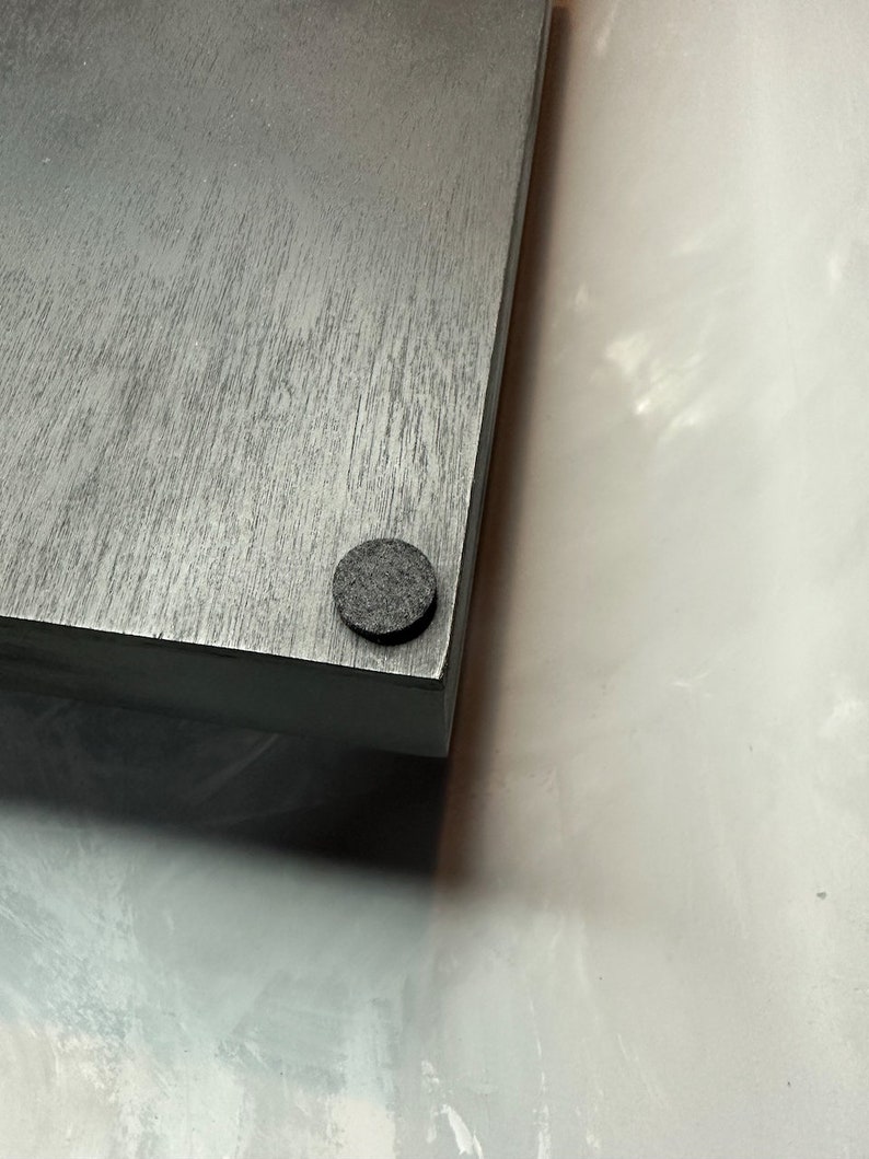 May include: A corner view of a gray wooden board with a dark gray felt pad attached to the bottom corner. The wood grain is visible on the surface of the board. The background is a light, textured surface.