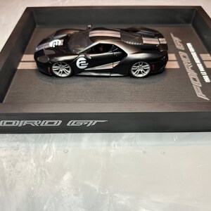 May include: A black Ford GT model car with white racing stripes and the number "2" on the side, displayed in a black wooden tray. The tray has the words "FORD GT" and "1968 LE MANS HERITAGE EDITION" printed on it.