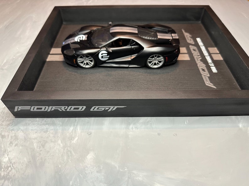 May include: A black Ford GT model car with white racing stripes and the number "2" on the side, displayed in a black wooden tray. The tray has the words "FORD GT" and "1968 LE MANS HERITAGE EDITION" printed on it.