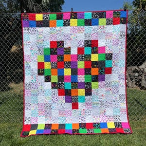 May include: A vibrant, handmade quilt featuring a pixelated heart design. The heart is made of colourful squares, surrounded by a white background with glasses patterns. The quilt has a red border and is hanging outdoors.