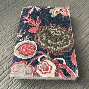 Puede incluir: Una funda de diario acolchada con un estampado floral. La funda presenta un fondo azul marino con un diseño repetido de flores y hojas rojas, blancas y verdes. Los bordes están cuidadosamente cosidos.