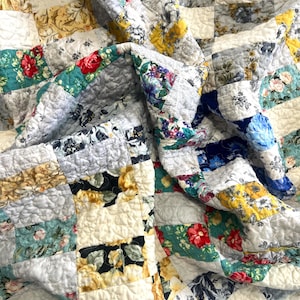 Puede incluir: Un edredón de retazos con una variedad de telas estampadas florales y con patrones en tonos de azul, amarillo, rojo y blanco. El edredón está hecho de cuadrados cosidos, creando una apariencia texturizada y acogedora. El edredón está doblado, mostrando los diferentes patrones.