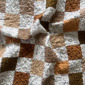 Puede incluir: Un edredón patchwork con cuadrados texturizados en tonos marrón, blanco y verde oliva. Algunos cuadrados presentan estampados florales, mientras que otros tienen un aspecto liso o acolchado. El edredón está doblado, mostrando su construcción en capas.