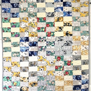 May include: A patchwork quilt with a variety of floral patterns in shades of blue, yellow, red, and green. The quilt is made of rectangular fabric pieces, with a gray border. The quilt is hanging on a white wall.