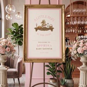 Könnte beinhalten: Ein gerahmtes Babyparty-Schild auf einer Staffelei. Das Schild hat einen goldenen Rahmen und zeigt den Text "A Little Bun in the Oven" mit einem Cartoon-Hasen als Koch. Der Text "Welcome to Sophia's Baby Shower" und das Datum 07.07.2026 sind ebenfalls enthalten.