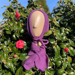 May include: A knitted purple hood and scarf set displayed on a mannequin head. The hood has a rounded shape with a tie closure, and the scarf features a leaf-like design. The set is made of a textured knit fabric.