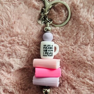 May include: A white ceramic mug keychain with the text "And here we go fucking again I mean good morning" stacked on top of three colourful silicone book charms. The keychain is attached to a silver metal ring.
