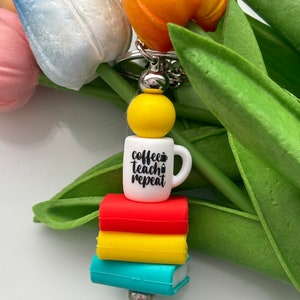 May include: White coffee mug keychain with the text "coffee teach repeat" and a pencil icon. The mug is stacked on top of three colourful books, red, yellow and blue.