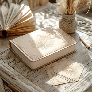 May include: A closed, cream-colored book with a gold embossed design on the cover. The book is sitting on a light wood table with a deck of tarot cards in front of it.