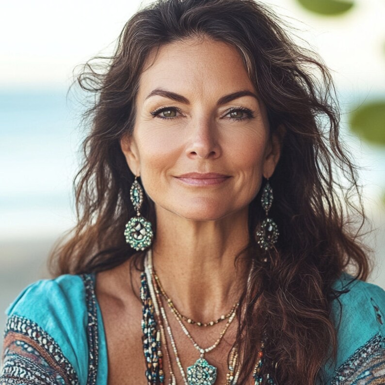 May include: A woman with long brown hair and green eyes is wearing turquoise earrings and a turquoise and gold necklace. She is also wearing a blue top with a patterned design. The background is blurred, suggesting an outdoor setting.