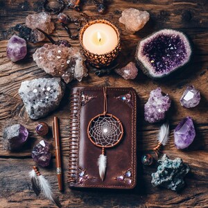 May include: A brown leather journal with a spiral binding and a dreamcatcher charm. The journal is surrounded by amethyst crystals, rose quartz, and other crystals. There is a lit candle and a pen on the table.