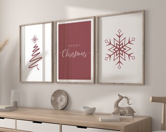 May include: Three framed Christmas-themed art prints. The prints feature a red Christmas tree, the words "Merry Christmas", and a snowflake design. The frames are a light brown color, and the prints are displayed on a light wood console table.
