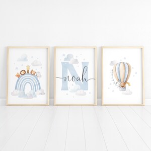 May include: Three framed art prints with a nursery theme. The prints feature a rainbow with animals, the name "Noah" with a blue "N", and a hot air balloon with a giraffe. The color palette includes blue, beige, and white.