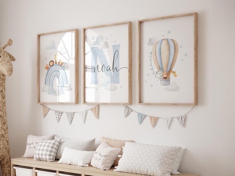 May include: Three framed art prints with a nursery theme. One print features a rainbow, another has the name "Noah", and the third shows a hot air balloon with a giraffe. The prints are in light wood frames and are hung above a bench with pillows.