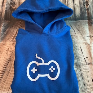 May include: A folded royal blue hoodie with a white controller graphic. The hoodie has a hood and is made of a soft material. The controller graphic is centered on the front of the hoodie.