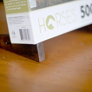 May include: A white puzzle box with the word "HORSES" and a horse head logo is propped up on a dark wooden stand. The box also has the number "500" printed on it. The stand is made of two pieces of wood.