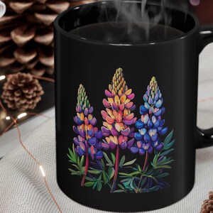 以下が含まれることがあります： 黒色のコーヒーカップに、紫、ピンク、青、黄色の花びらを持つルピナスが3つ描かれたカラフルな花柄のデザインが施されています。花は黒い背景に描かれています。