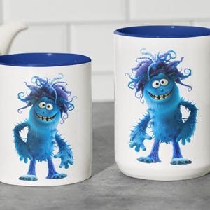 May include: Two white ceramic mugs with blue rims. Both mugs feature a cartoon blue monster with a toothy grin and wild blue hair. The monster is standing with its arms at its sides.