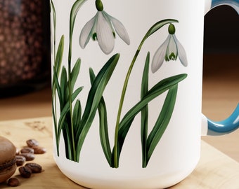 May include: White ceramic mug with a blue rim and handle. The mug features a botanical illustration of three white snowdrop flowers with green leaves.