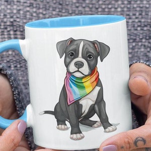 May include: A white ceramic mug with a light blue rim and handle. The mug features a cartoon illustration of a black and white puppy wearing a rainbow bandana.
