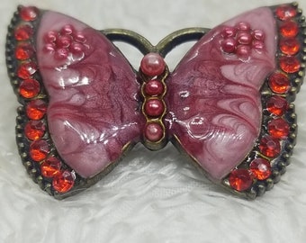May include: A pink and white butterfly brooch with red rhinestone accents. The butterfly has a marbled design and is set in a bronze-colored metal.