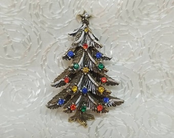 May include: A gold-tone Christmas tree brooch with white enamel and colorful rhinestones. The tree is decorated with red, blue, green, and yellow rhinestones.