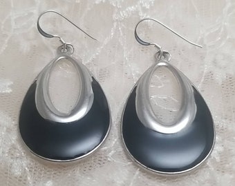 May include: A pair of silver dangle earrings with black enamel. The earrings are shaped like teardrops with a silver oval cutout in the center.