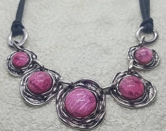 May include: A silver necklace with a black cord and five pink, round, textured stones. The stones are set in silver circles.