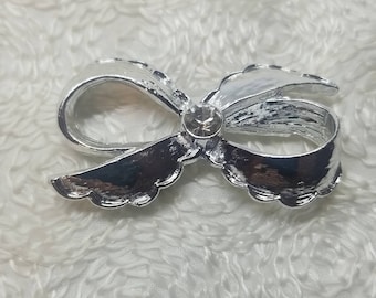 May include: Silver bow brooch with a clear rhinestone in the center. The bow has a scalloped edge.