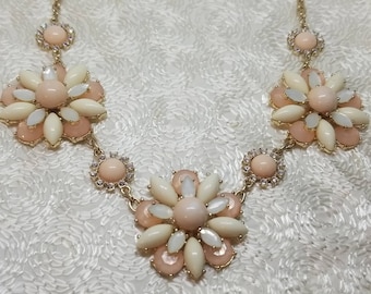 May include: A gold-toned necklace featuring large, flower-shaped pendants. Each flower has a central peach-colored stone surrounded by cream and peach-colored petals. Smaller, circular accents with clear stones and peach centers connect the flowers.