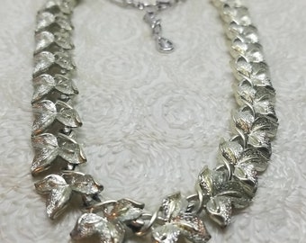 May include: A silver tone necklace with a leaf design. The necklace has a clasp closure and is made of metal.