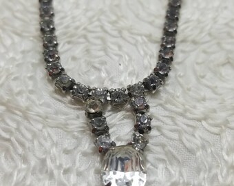 May include: A silver necklace with a large, clear rhinestone pendant. The necklace is made of small, clear rhinestones.
