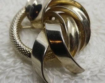May include: A vintage silver-toned brooch with a textured, circular frame and a swirling, ribbon-like design. The brooch has a polished finish and a pin closure on the back. The brooch is a classic accessory.