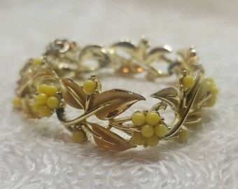 May include: A gold-tone bracelet with a delicate design of leaves and yellow flowers. The bracelet is made of metal and has a vintage look.