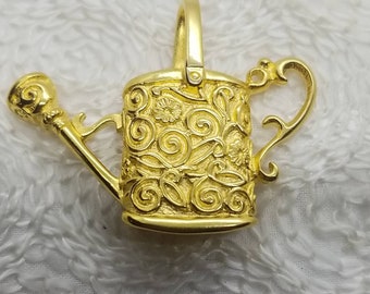 May include: A gold-colored metal watering can charm with a detailed floral design. The charm has a loop at the top for attaching to a necklace or bracelet.