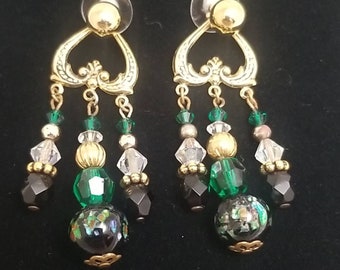May include: Gold tone dangle earrings with black, green, and clear crystal beads. The earrings have a heart-shaped top with a gold tone design.