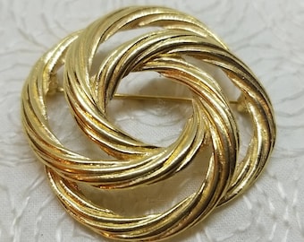 May include: A gold-toned metal brooch in the shape of a knot. The knot is made of multiple intertwined strands of metal, creating a circular design.