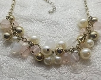 May include: A gold chain necklace with white and pink beads. The necklace has a variety of different sized beads, including pearls, glass beads, and metal beads.
