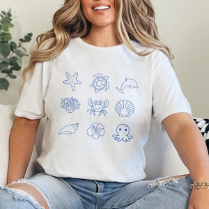 Puede incluir: Una camiseta blanca con un contorno azul de varias criaturas marinas, incluyendo una estrella de mar, una tortuga, un delfín, un coral, un cangrejo, una concha, una concha, una flor de hibisco y un pulpo.