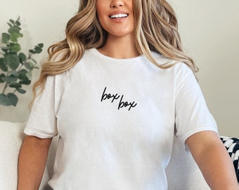 Puede incluir: Una camiseta blanca con el texto "box box" escrito en cursiva negra. La persona que lleva la camiseta está sentada en un sofá blanco con una manta de cuadros negros y blancos.