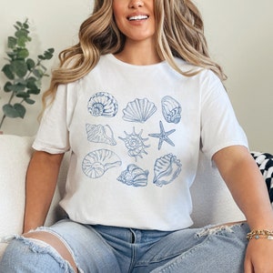 Könnte beinhalten: Ein weißes T-Shirt mit einem blauen Muschel-Design. Das Design zeigt eine Vielzahl von Muscheln, darunter einen Seestern, eine Nautilus-Schale und eine Venusmuschel.
