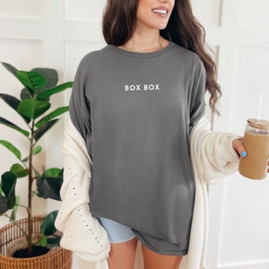 May include: A woman wearing a gray long-sleeved shirt with the text "BOX BOX" printed on the front. She is wearing a white cardigan and blue denim shorts. She is holding a glass with a brown beverage.